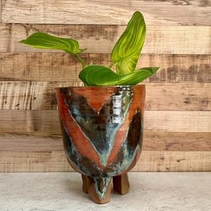 May include: A ceramic planter with three short legs featuring a glossy glaze in shades of burnt orange, deep brown, and teal. The pot contains a vibrant green plant with broad, pointed leaves that have prominent light green veins throughout.