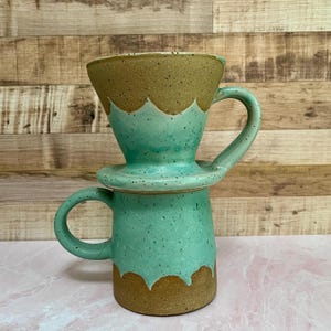 May include: A handmade ceramic coffee pour-over set. The set includes a turquoise mug with a handle and a matching pour-over cone. The top of the cone and the bottom of the mug have a brown, speckled finish. The set is designed for brewing coffee.