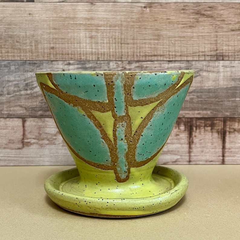 May include: A handmade ceramic planter with a matching saucer. The planter is cone-shaped, with a light green and yellow color scheme. The design features a geometric pattern in brown. The saucer is round and yellow. The planter is suitable for small plants.