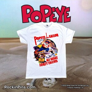 May include: White t-shirt with a vintage illustration of Popeye the Sailor, holding a pickaxe and standing on a pile of treasure chests. The text "Popeye the Sailor Meets Ali Baba's Forty Thieves" is printed on the shirt.