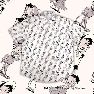 May include: A white short-sleeved button-up shirt with a repeating pattern of Betty Boop in various poses. The shirt has a collar and a pocket on the left chest. The text "TM & © 2023 Fleischer Studios" is printed at the bottom of the shirt.