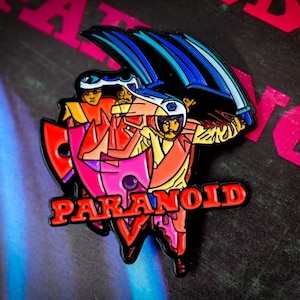 May include: A colorful enamel pin featuring a stylized illustration of two figures in space helmets and armor, with a sword and shield. The word "PARANOID" is in red, with a pink and red shield shape. The background is dark.