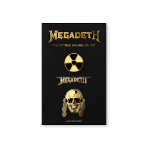 May include: A collectible enamel pin set featuring the band Megadeth. The set includes a gold and black radiation symbol pin, a gold band logo pin, and a gold skull pin with sunglasses and chains. The pins are displayed on a black card.