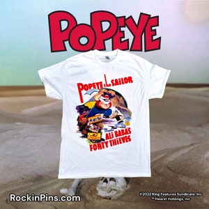 May include: White t-shirt featuring a graphic of Popeye the Sailor, with the text "Popeye the Sailor Meets Ali Baba's Forty Thieves." The shirt has a crew neck and short sleeves. The Popeye logo is in red. RockinPins.com is printed at the bottom.