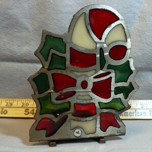 May include: A stained glass ornament featuring a candy cane, holly leaves, and a bow in red, green, and off-white colors, framed in metal. A yellow measuring tape is visible behind the object, showing a portion of the scale in inches.