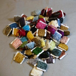 May include: A collection of colorful embroidery floss spools, each wrapped in a small cardboard box with a label. The spools are arranged in a pile, showcasing a variety of shades and textures.