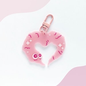 May include: A pink worm-shaped keychain with a rose gold clasp. The worm has a heart-shaped opening and is decorated with small stars and wavy lines. The keychain is on a white and pink background.