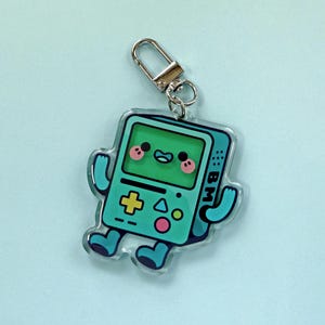 May include: A clear acrylic keychain featuring a cartoon character shaped like a teal handheld gaming console with a green screen, pink cheeks, a yellow plus sign, and blue limbs. The device has the letters BM printed on the side and a silver clasp.