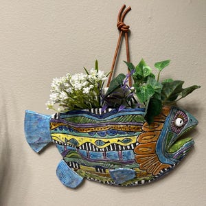 May include: A ceramic fish-shaped wall planter featuring a vibrant, multi-colored pattern of blue, yellow, green, and purple geometric shapes and small fish motifs. The planter is suspended by a brown leather cord and contains artificial greenery.