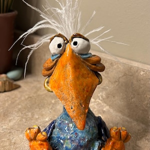 May include: A ceramic bird figurine with a large orange beak, wide white eyes, and wild white feathers on its head. The body is textured blue, and the feet are orange. The bird is in a seated position with its feet extended forward.
