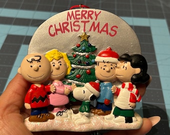 May include: A Christmas ornament featuring the Peanuts gang. The ornament has a silver background with the words "Merry Christmas" in red. The characters are in festive attire, with a Christmas tree in the center.