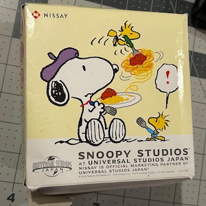 May include: A box featuring Snoopy and Woodstock enjoying spaghetti. Snoopy, wearing a purple beret, is seated and eating from a plate. Woodstock is also eating spaghetti. The box has the text "Snoopy Studios at Universal Studios Japan."