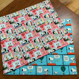 May include: Two fabric panels featuring a repeating pattern of Peanuts characters, including Snoopy and Woodstock. The top panel is pink with red hearts and the bottom panel is blue with white text that reads "OK, FULL POWER!" and "FASTER! FASTER! FASTER!"