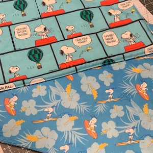 May include: A blue fabric with a repeating pattern of Snoopy riding a surfboard with hibiscus flowers and palm leaves. The fabric is folded over a second fabric with a comic strip pattern of Snoopy flying a hot air balloon and saying "Faster! Faster! Faster!" and "Okay, full power!"