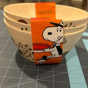 May include: A stack of three beige bowls with a cartoon illustration of Snoopy on an orange band. The bowls have a fall-themed design with leaves and other seasonal elements. The bowls are made of a durable material.