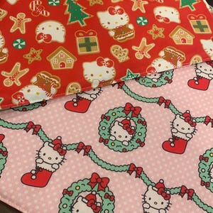 May include: Two fabric squares with a Christmas theme. The top square is red with a white polka dot pattern and features a repeated image of Hello Kitty dressed in a gingerbread costume. The bottom square is pink with a white polka dot pattern and features a repeated image of Hello Kitty wearing a Santa hat and holding a wreath.