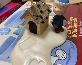 May include: A white ceramic Christmas ornament featuring Charlie Brown from Peanuts holding a wreath. The ornament also includes a small yellow dog house with a brown roof and a string of Christmas lights. The ornament is sitting on a white base with a blue and white sticker that says "Schultz".