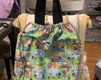 May include: A green drawstring bag with a Peanuts cartoon print featuring Snoopy, Charlie Brown, and other characters. The bag has black handles and pink and green drawstrings.