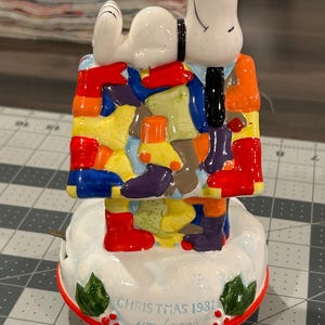 May include: A ceramic Snoopy figurine in a colorful house, with the text "CHRISTMAS 1981" and the serial number 4156/15000. The figurine features Snoopy in a black tie and colorful boots. The base is white with green holly.