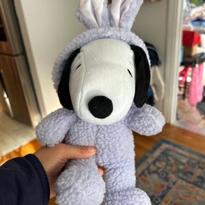 May include: A plush Snoopy toy wearing a light purple bunny costume. The toy has a white face, black ears, and a black nose. The bunny costume includes a hood with upright ears and a matching body.