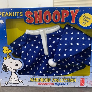 May include: A blue and white Peanuts Snoopy wardrobe collection nightshirt for a plush toy. The nightshirt has a white trim and a white heart pattern. The packaging features the Peanuts logo and the text "Collect Them All".