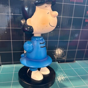May include: A figurine of Lucy from the Peanuts comic strip. The figure has black hair, a blue dress, and white shoes. The figurine is standing on a black base. The background is a grid pattern with blue and white squares.
