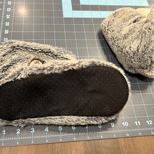 May include: Gray faux fur slipper boots with black non-slip soles. The boots have a plush, furry texture and a small wooden button detail. The soles are textured for grip. These cozy slippers are designed for indoor wear.