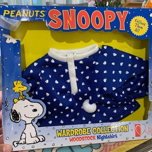 May include: A blue and white nightshirt with a white collar and button closure. The nightshirt has a white paw print pattern and says "Woodstock Nightshirt" on the packaging. The packaging also features a cartoon image of Snoopy and Woodstock.