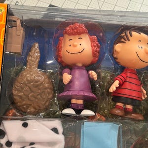 May include: A boxed set of Peanuts character figurines. The set includes figures of Lucy in a purple dress, Linus in a red and black striped shirt, and other themed accessories. The packaging is orange and clear plastic.