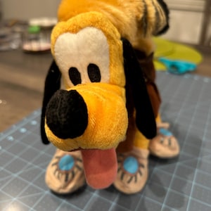 May include: A plush toy of Pluto, a cartoon dog character. The toy has a yellow head and body, black ears, and a pink tongue sticking out. The dog is wearing brown shorts and tan shoes with blue accents.