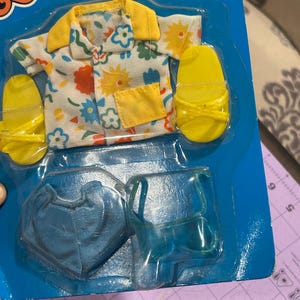 May include: A doll-sized outfit set, including a floral print shirt with yellow trim, matching yellow sandals, and blue accessories, all packaged on a blue cardboard backing. The shirt features a colorful floral pattern. The packaging reads "Fun Fas" and "Collection".