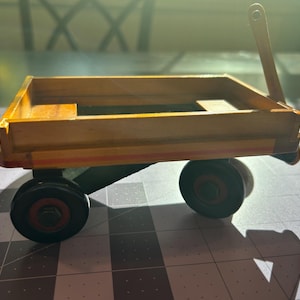 May include: A small wooden toy wagon with a pull handle. The wagon bed is rectangular and painted in a light brown color with red and yellow stripes. The wheels are black with red accents.