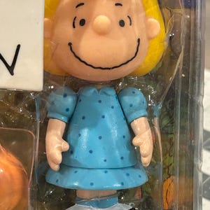 May include: A figurine of a cartoon character with blonde hair and a blue bow. The character is wearing a light blue dress with dark blue polka dots and white shoes. The figurine is in a clear plastic package.