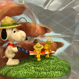 May include: A Snoopy figurine wearing a khaki hat and red scarf, holding a miniature American flag with Woodstock and two other yellow birds. The figurine is on a green grassy base.