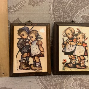 May include: Two framed Hummel prints. The prints feature children in vintage clothing. The frames are dark brown with a metal loop for hanging. The artwork is in muted tones of beige, blue, and brown. The prints are approximately 5 inches tall.