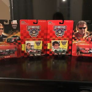 May include: Four unopened boxes of NASCAR 50th Anniversary die-cast cars. The boxes feature red, white, and blue color schemes with the NASCAR 50th Anniversary logo. Each box includes a die-cast car and a photo of a NASCAR driver.