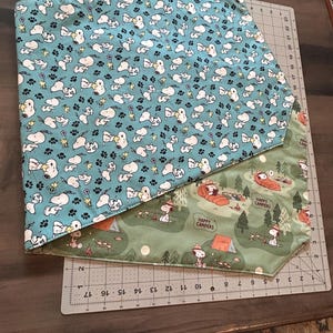 May include: Reversible dog bandana featuring Snoopy and paw prints on a teal background, and a camping scene with Snoopy on a green background. The bandana is made of fabric and is likely handmade.