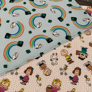 May include: Two fabric squares with different patterns. The top square is light blue with white polka dots and a repeating pattern of rainbows and pots of gold. The bottom square is white with light purple polka dots and a repeating pattern of Peanuts characters.
