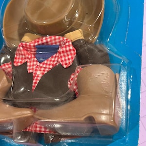 May include: A cowboy doll outfit in a plastic package. The outfit includes a brown cowboy hat, a brown jacket with a red and white checkered shirt, and brown cowboy boots. The packaging is blue.