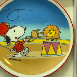 May include: A round ceramic plate featuring a colorful illustration of Snoopy dressed as a circus ringmaster holding a chair toward Woodstock, who is styled as a lion inside a circus ring. The name Schulz is printed in black on the lower left.