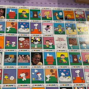 May include: A large rectangular sheet featuring a grid of colorful Peanuts comic strip panels, including illustrations of Snoopy, Charlie Brown, and other characters, alongside two photographic portraits of Charles M. Schulz, with a ruler visible at the top.