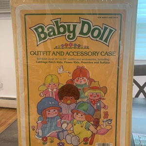 May include: A yellow and brown cardboard storage case with a green handle. The front of the case has a picture of a group of baby dolls and the text "Baby Doll Outfit and Accessory Case". The text "Will hold most 16" to 18" outfits and accessories, including Cabbage Patch Kids, Flower Kids, Preemies and Softina" is printed below the picture. The case is labeled "No. 11300".