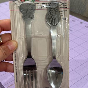 May include: A vintage stainless steel baby fork and spoon set featuring Snoopy from the Peanuts comic strip. The fork and spoon have embossed Snoopy designs on the handles and are packaged in a clear plastic blister pack.