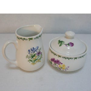 Puede incluir: Un juego de jarra de crema y azucarero de cerámica blanca con un diseño floral. La jarra tiene un asa y el azucarero tiene una tapa. Ambas piezas presentan un borde de vid verde y flores de colores.