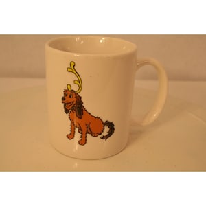 May include: White ceramic mug with a cartoon illustration of a brown dog wearing a yellow reindeer antler headband.
