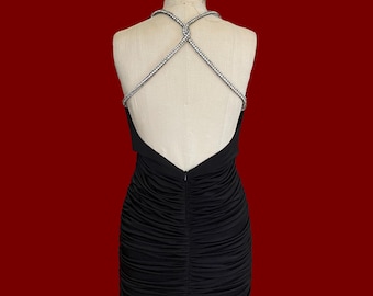 May include: Black mini dress with a low back and rhinestone straps. The dress features a ruched design on the front and back. The dress is displayed on a mannequin against a red background.