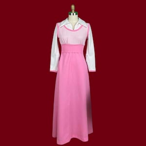Puede incluir: Un vestido largo rosa vintage con una camisa blanca con cuello y mangas largas. El vestido presenta un corpiño rosa texturizado y una falda rosa a juego. El vestido se exhibe en un maniquí sobre un fondo rojo.