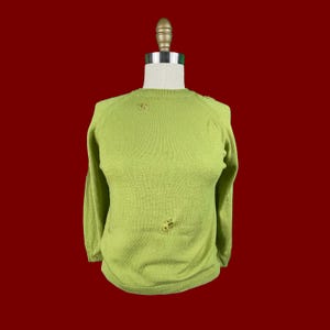 Op de afbeelding: Een lichtgroene trui met lange mouwen en een ronde hals, tentoongesteld op een mannequin. De trui heeft kleine, decoratieve versieringen en een ietwat versleten uiterlijk. De achtergrond is effen rood.