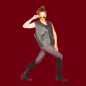 May include: A black leather vest with a silver zipper is worn over a black top. The person is wearing heart-shaped yellow sunglasses, dark tights, and black knee-high boots with zippers. The background is a solid dark red.
