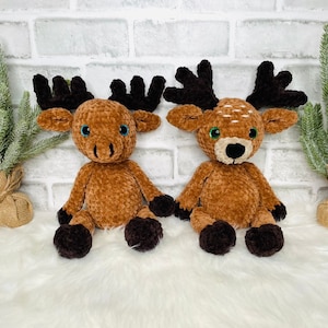 May include: Two plush crocheted stuffed animals, a brown moose and a brown deer, sitting on a white fluffy surface. Both animals have brown eyes and black noses.